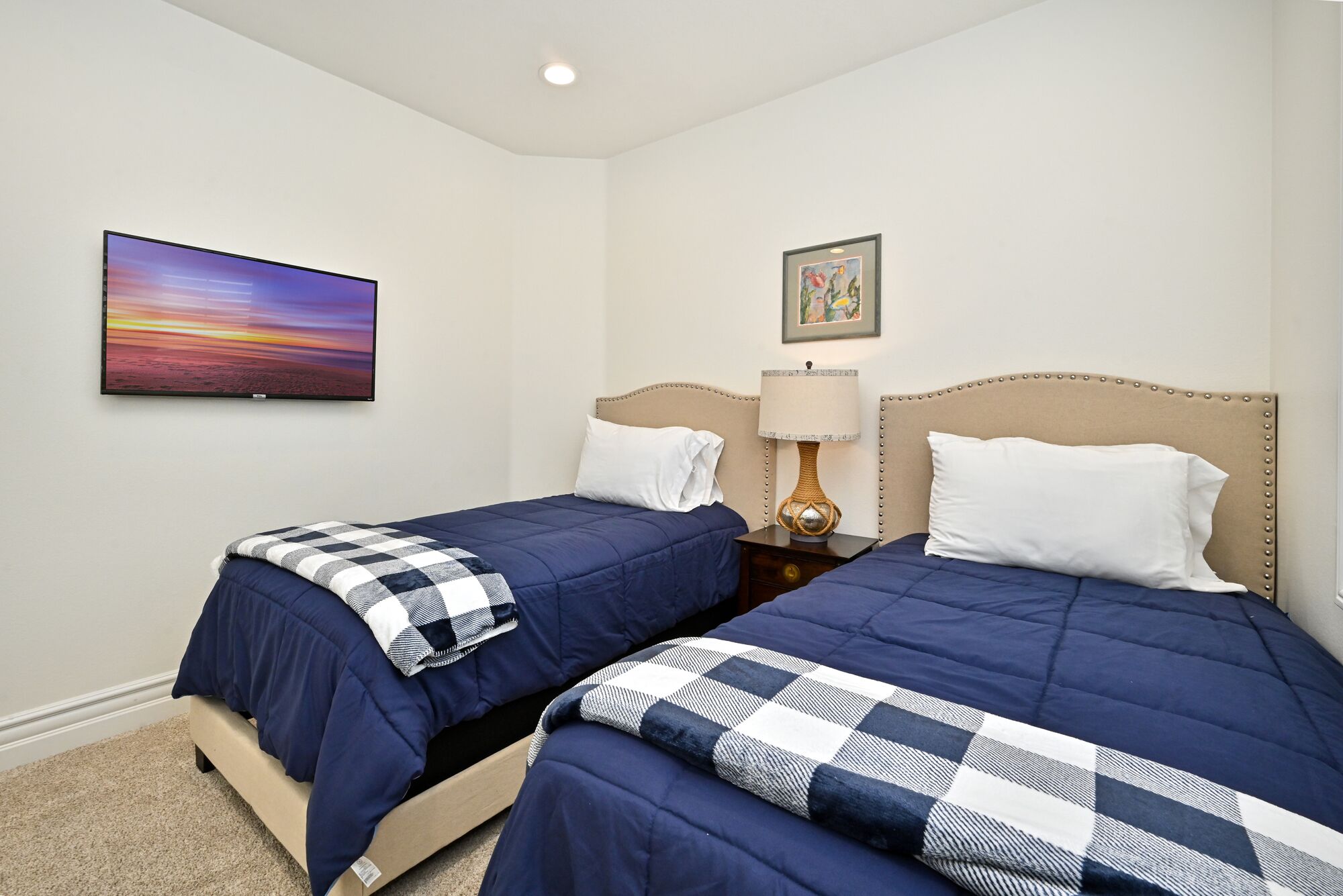 A hotel room with two beds, a painting on the wall, and a lamp on the nightstand.