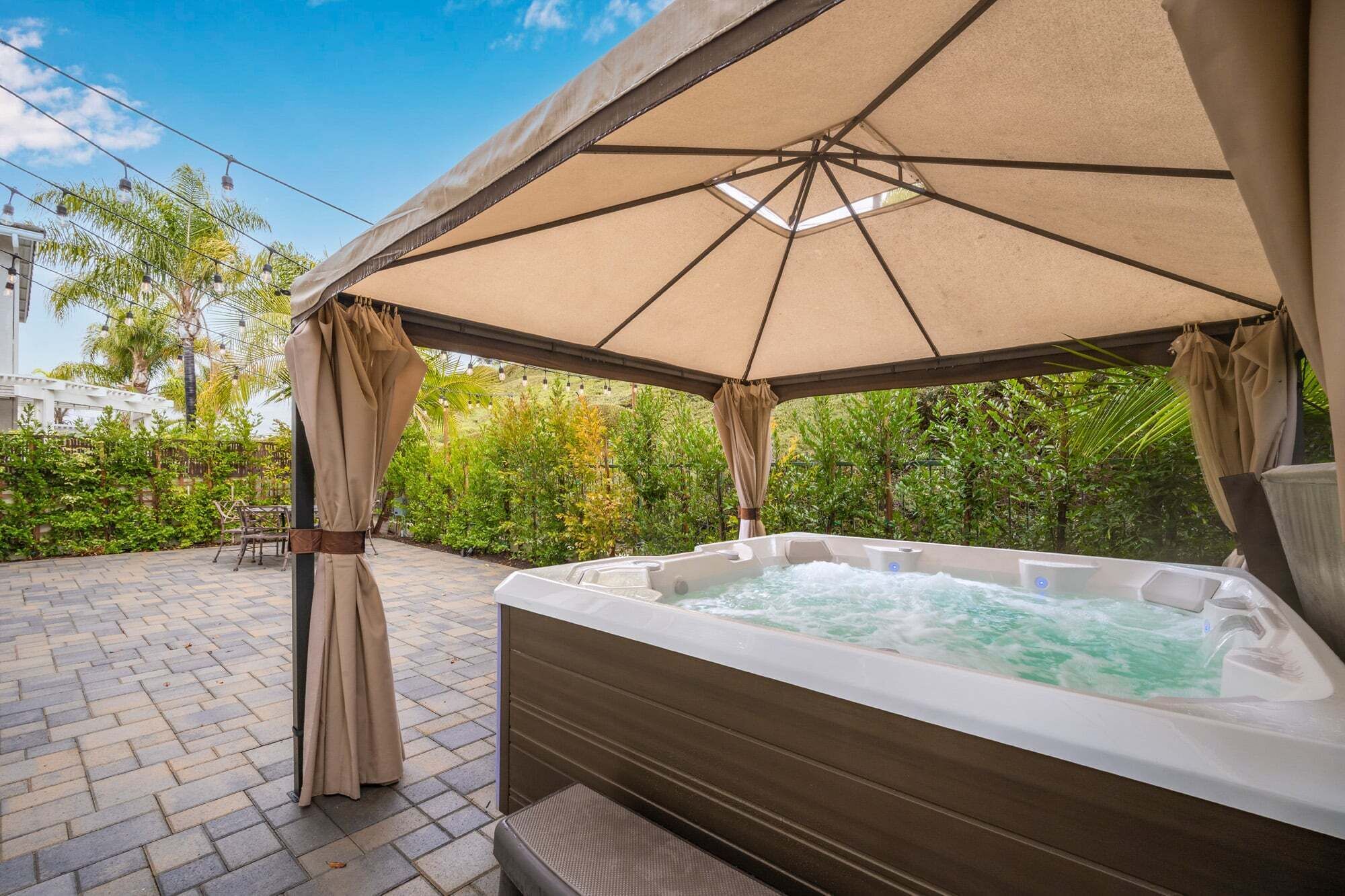 A hot tub under a canopy with a brick patio and lush greenery in the background.