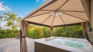 Un jacuzzi bajo una pérgola, con un patio de ladrillo y abundante vegetación al fondo.