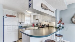 A modern kitchen with white cabinets, a large island with stools, a refrigerator, and a microwave. - Ground floor | Beachfront Corner Unit a few Steps from the Beach (Fernandina Beach)