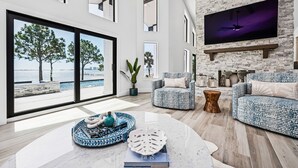 A modern living room with a large window overlooking a beach, a fireplace, and a flat-screen TV mounted on a brick wall.