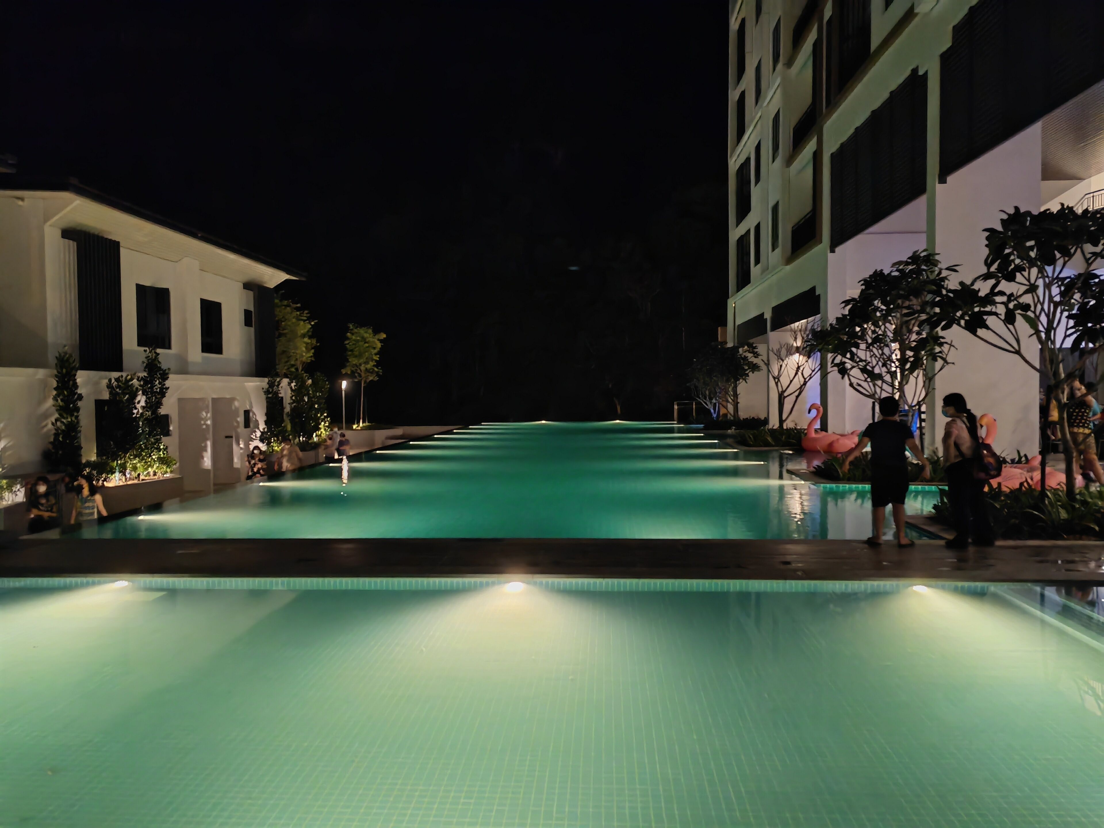 A nighttime swimming pool with a clear water surface, surrounded by buildings and trees.
