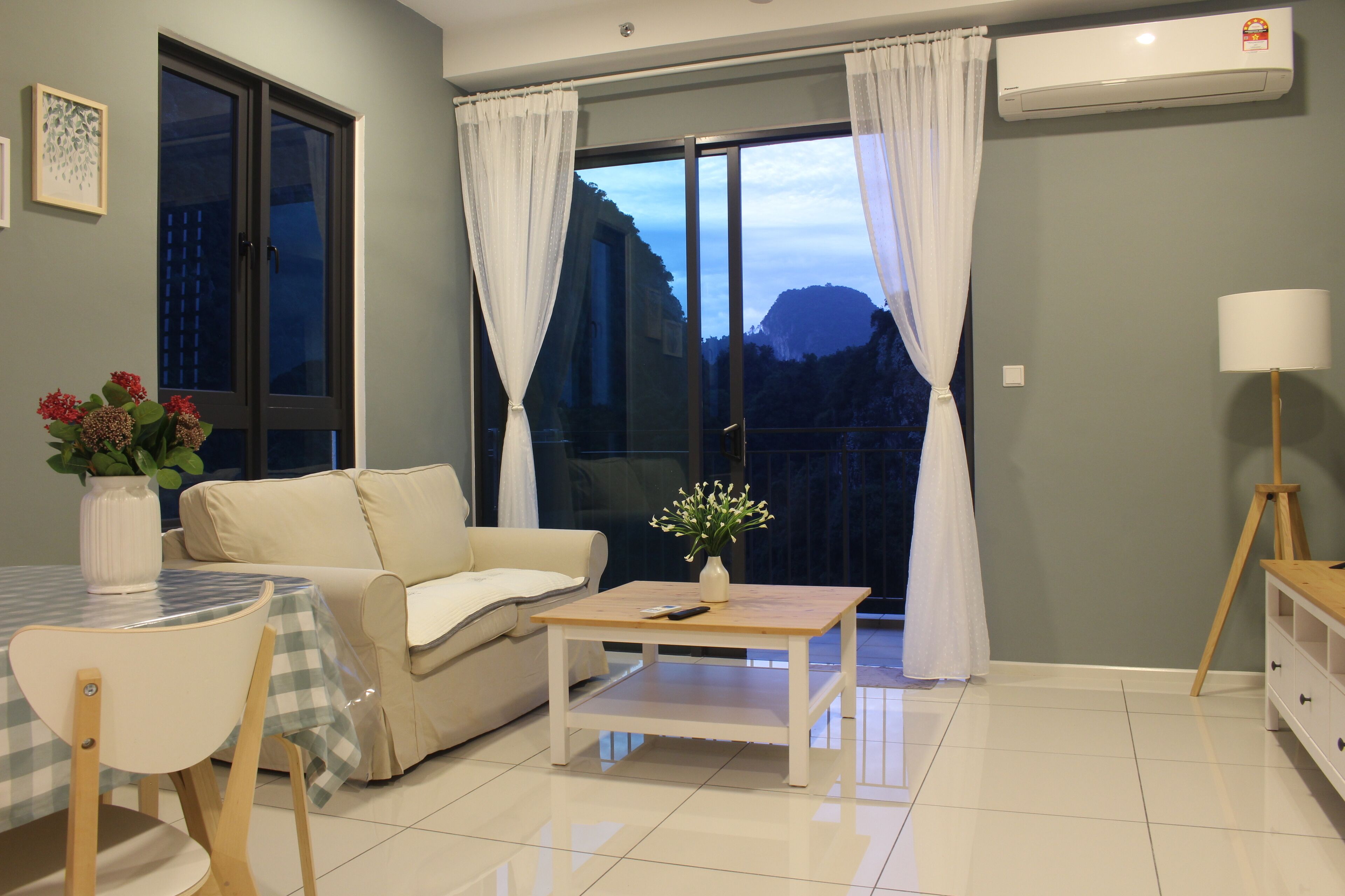 A living room with a sofa, a table, and a chair. There is a window with curtains, a vase with flowers, and a lamp.