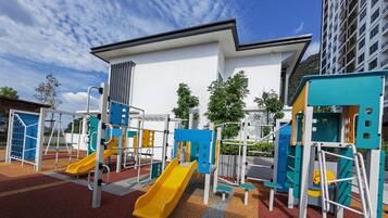 A playground with slides and climbing structures, surrounded by a building with a white facade and a red-tiled ground.