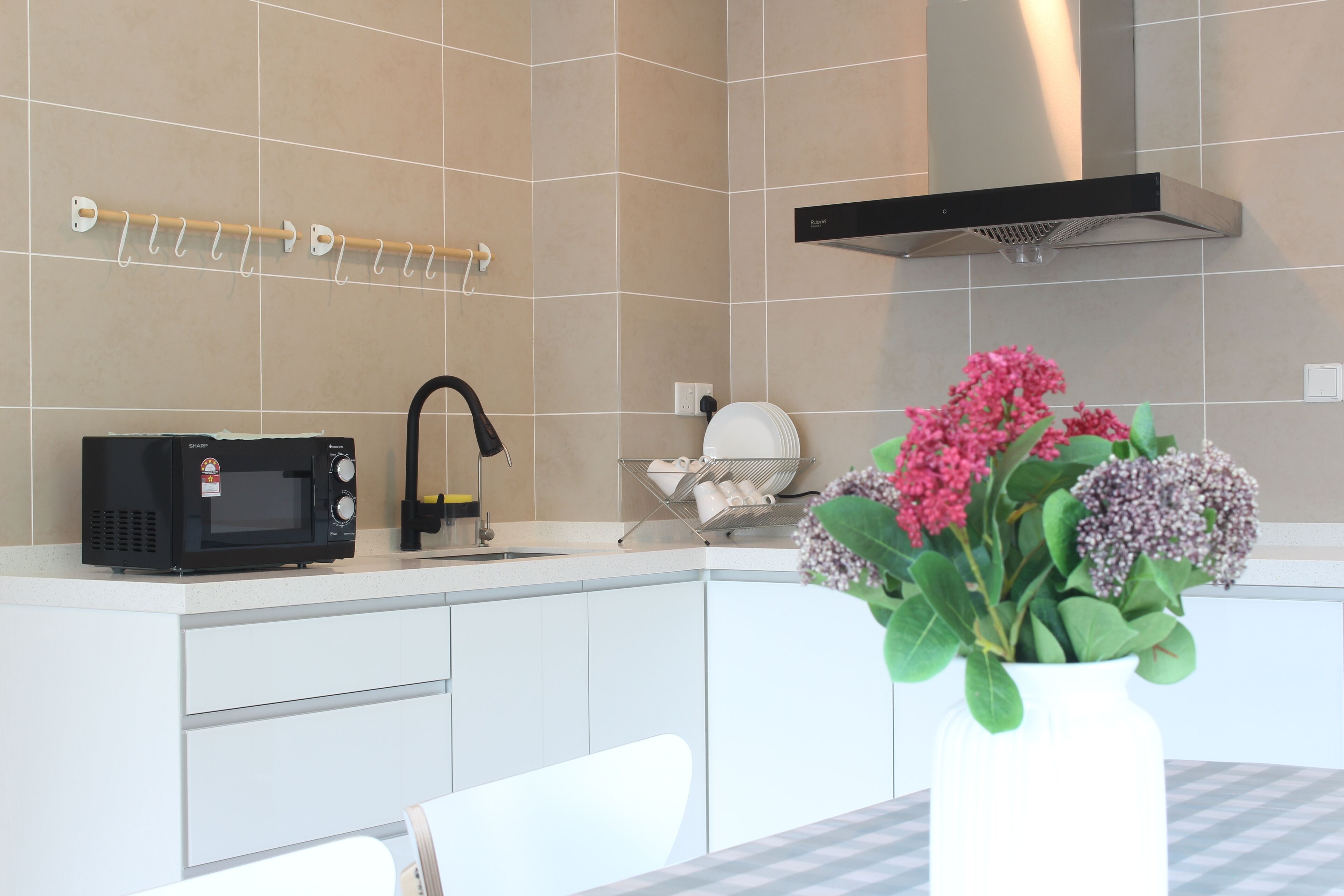 A modern kitchen with white cabinets, a microwave, a sink, and a stainless steel range hood. There is a white vase with flowers on the counter.
