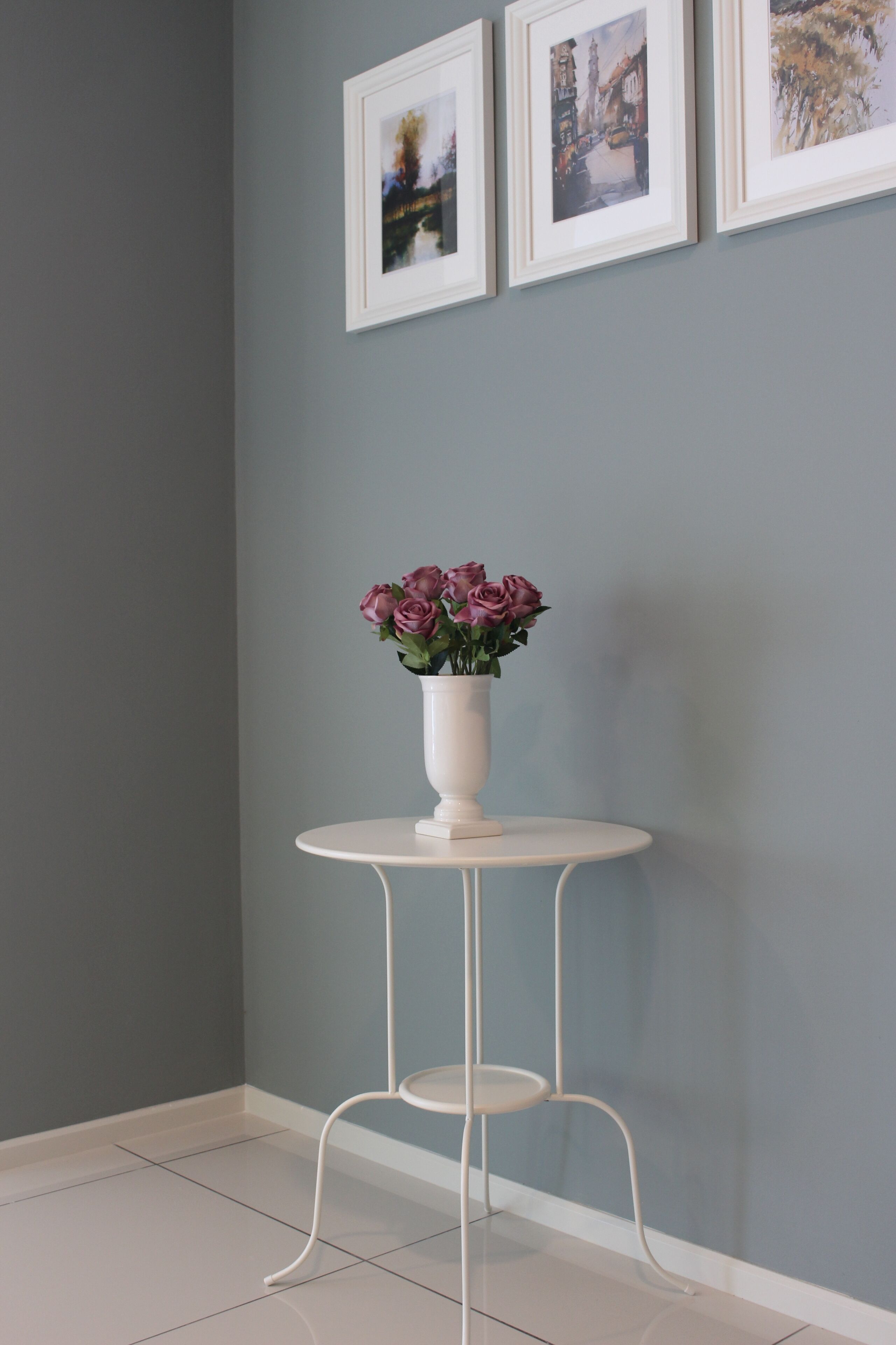 Quarto de canto com mesa branca redonda e cadeira branca, um vaso com flores rosa, e três quadros na parede.