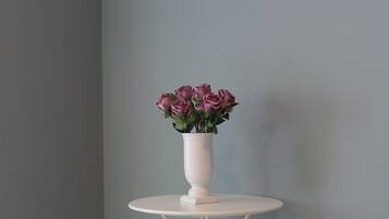 Quarto de canto com mesa branca redonda e cadeira branca, um vaso com flores rosa, e três quadros na parede.