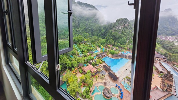Vista desde una ventana con vistas a un complejo turístico, un área de piscina, abundante vegetación y terreno montañoso.