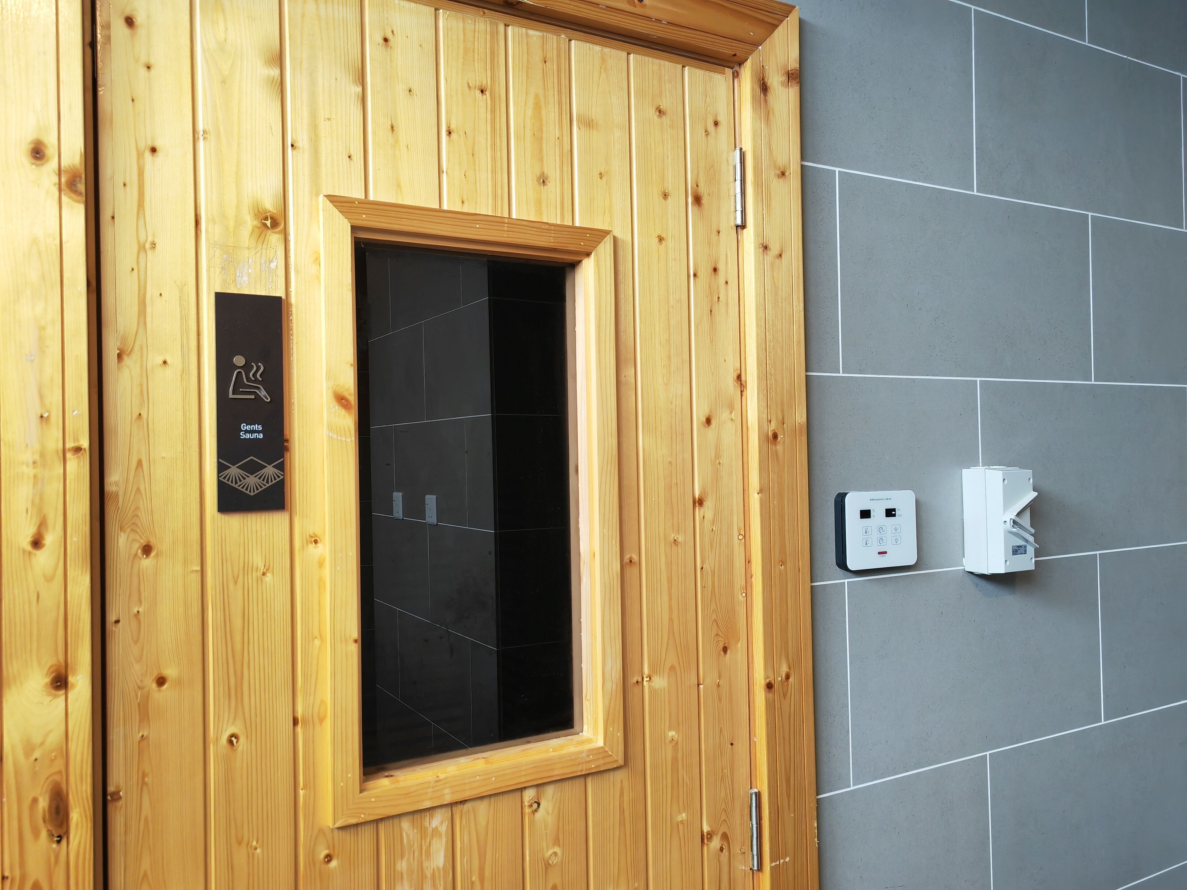 Een houten sauna deur met een zwart glasraam, een bedieningspaneel en elektrische stopcontacten op een betegelde muur.
