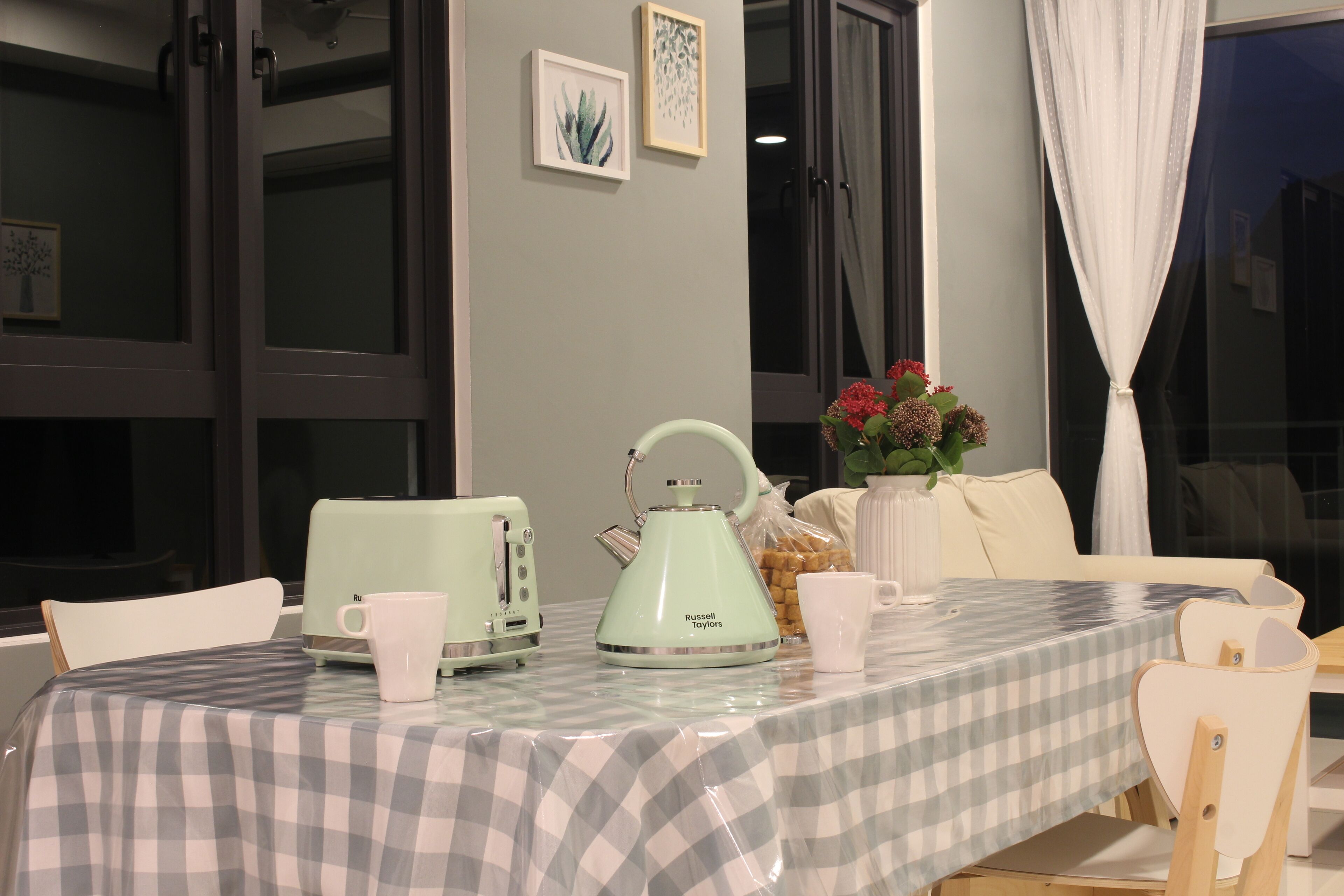 A dining area with a checkered tablecloth, a green kettle, a white mug, and a vase with red flowers.