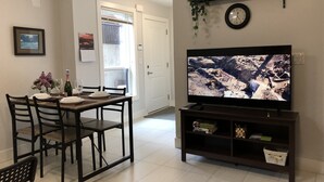 A living room with a dining table set for four, a television on a stand, and a door leading to a balcony. - Beautiful 5 star 2 bd suite near bus&shopping (Burnaby)
