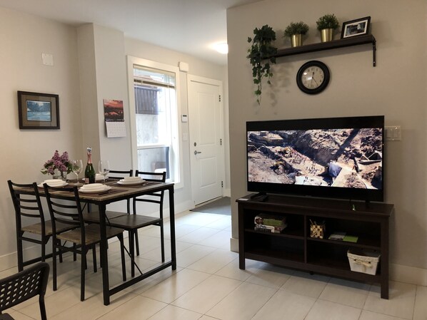 A living room with a dining table set for four, a television on a stand, and a door leading to a balcony.