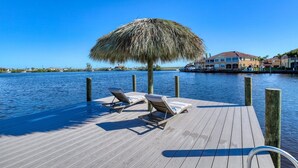 A wooden dock with lounge chairs and a thatched umbrella, overlooking a calm body of water with buildings in the background.