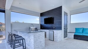 An outdoor kitchen with a bar area, a TV mounted on a wooden wall, and a blue sofa.