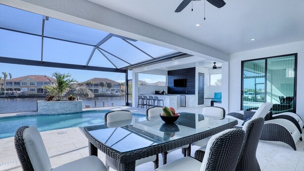 A modern outdoor dining area with a glass table and wicker chairs, overlooking a swimming pool and a marina.