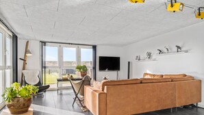 A modern living room with a large sofa, a TV mounted on the wall, and a large window offering a view of the outdoors. - 3 bedroom nice home in Thyborøn (Thyborøn)