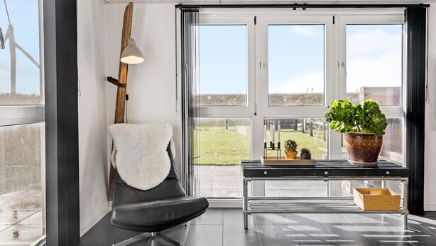A modern room with a large window, a black chair with a white throw, a table with a potted plant, and a candle holder. - 3 bedroom nice home in Thyborøn (Thyborøn)