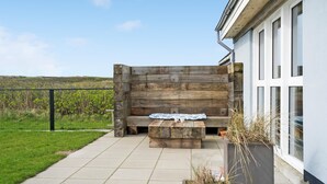 An outdoor seating area with a wooden bench and a table, a concrete pathway, and a view of a grassy field and fence. - 3 bedroom nice home in Thyborøn (Thyborøn)