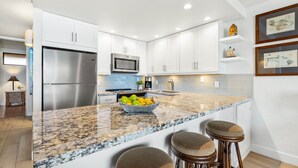 A modern kitchen with white cabinets, stainless steel appliances, a granite countertop, and a bowl of fruit. - Stunning oceanfront view with Maui-inspired design (Lahaina)