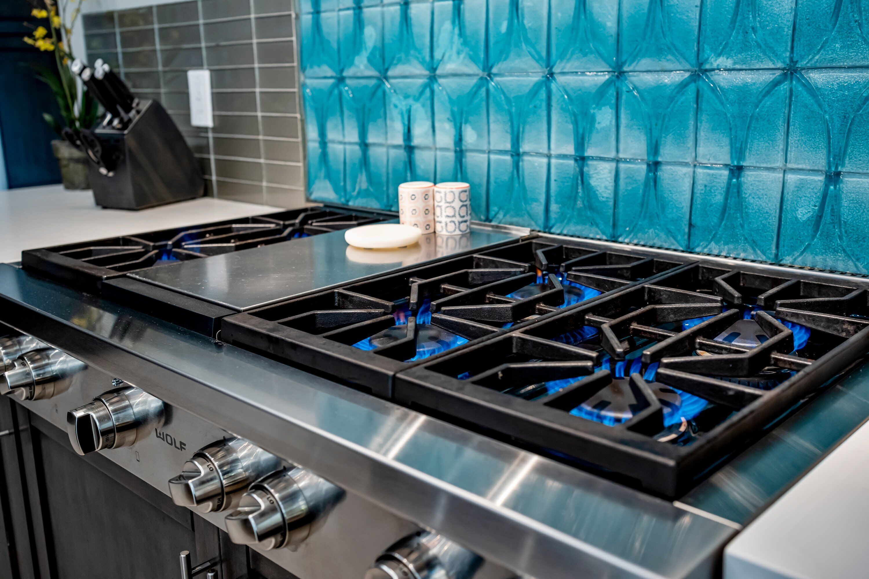 Une cuisine équipée d’une cuisinière Wolf en acier inoxydable, d’un plan de travail blanc et d’un dosseret de carrelage bleu.