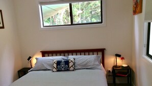 A neatly made bed with a headboard, two bedside tables with lamps, a window with a view of trees, and a painting on the wall.