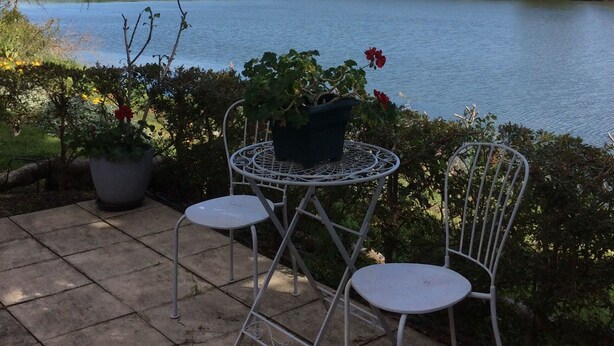 A patio with two white metal chairs and a table, overlooking a lake with mountains in the background.