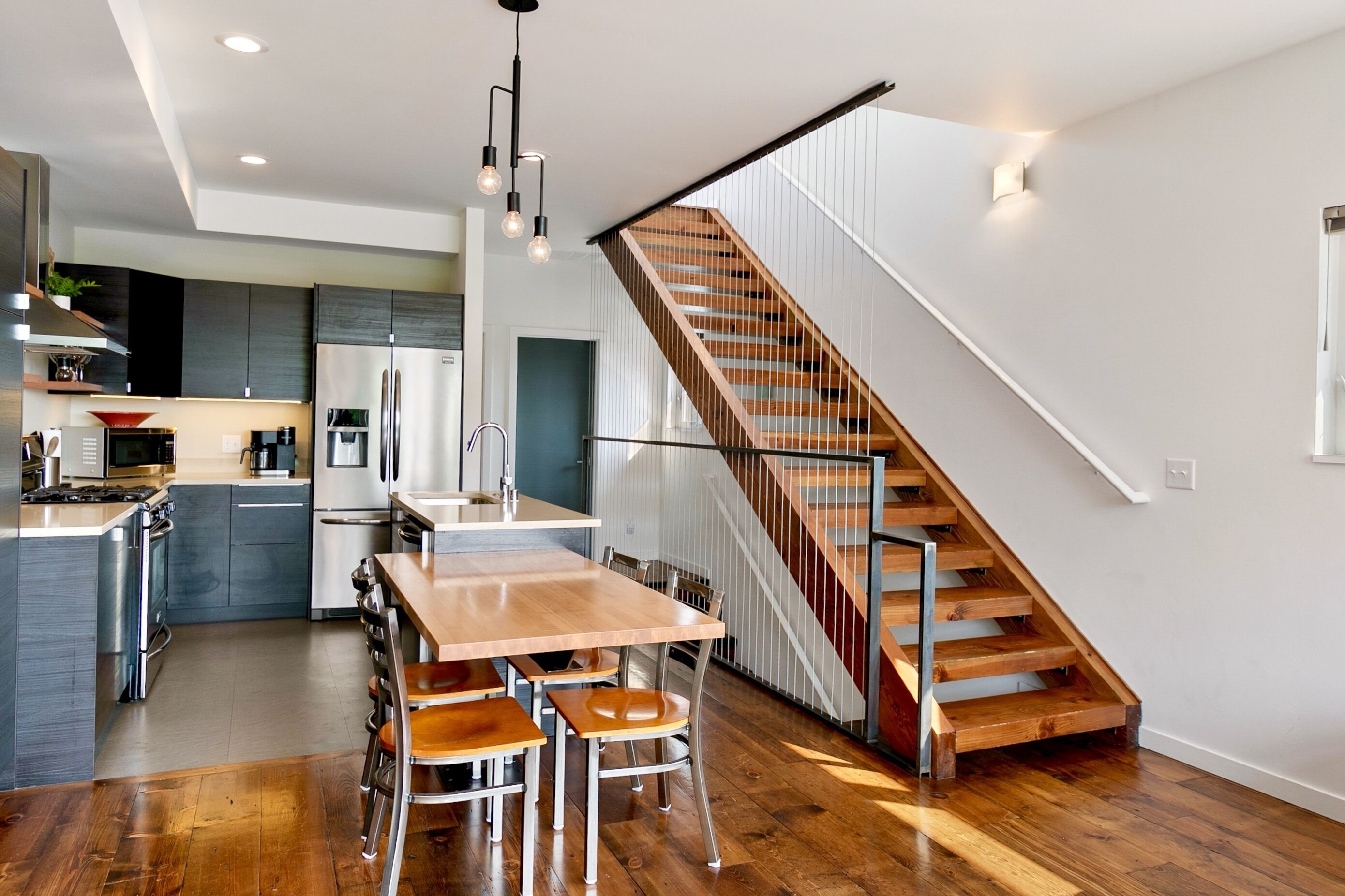 A modern kitchen with a wooden dining table and chairs, a stainless steel refrigerator, and a wooden staircase.