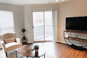 Una sala de estar con un sofá, una mesa de centro, un televisor sobre un mueble bajo y un ventanal corredizo que da a un espacio exterior.