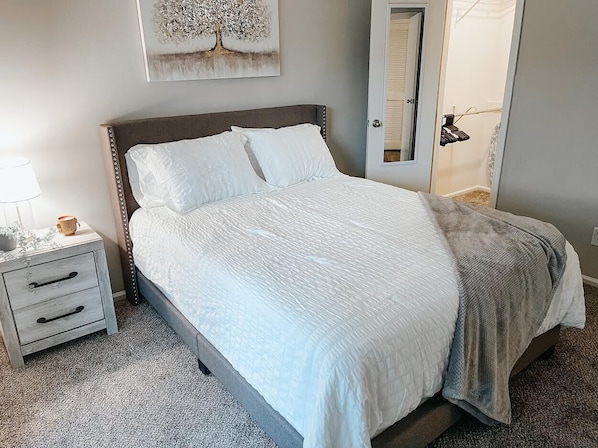 Una cama bien hecha con sábanas blancas, cabecera de madera y una manta gris.