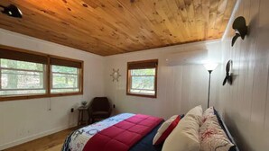 Quarto com teto de madeira, duas janelas, uma cama com colcha vermelha e azul, e uma mesa de cabeceira.