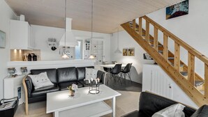 Un salon moderne avec un canapé en cuir noir, une table basse blanche et un coin repas avec des chaises noires. On aperçoit un escalier en bois à l’arrière-plan.
