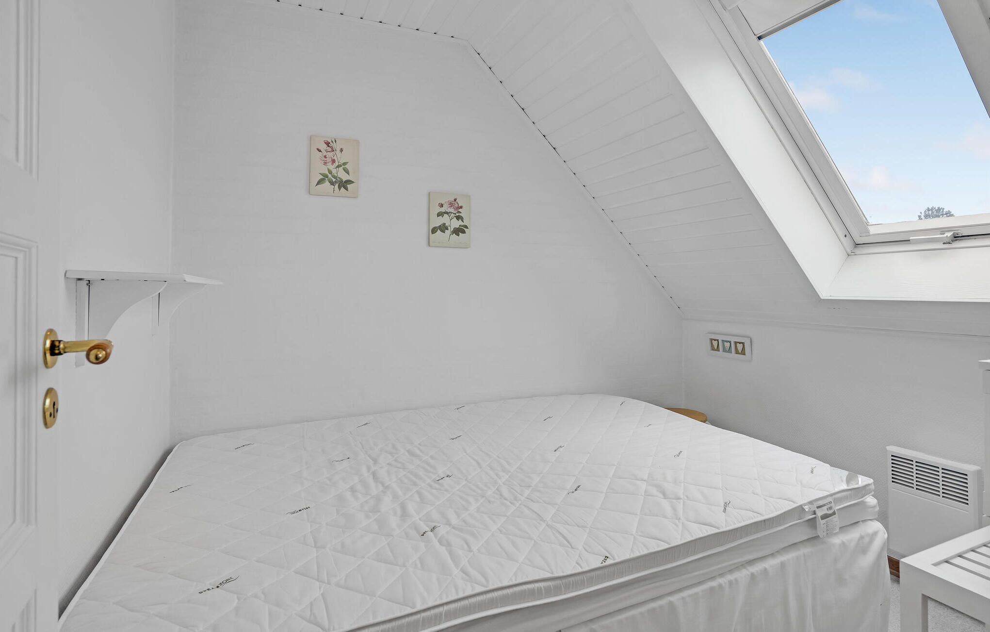 A bedroom with a sloped ceiling, a skylight, a bed with a white quilted mattress, and two wall-mounted floral paintings.