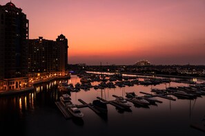 Un puerto deportivo al atardecer con barcos amarrados, un horizonte urbano de fondo y edificios con ventanas iluminadas.