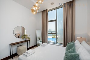 Una habitación de hotel moderna con una cama grande, una mesita de noche, un espejo y vistas a una playa y al paisaje urbano a través de un ventanal amplio.