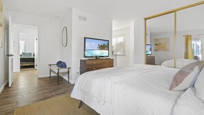 Un dormitorio con una cama grande, un televisor sobre un aparador de madera, un espejo y vistas a una playa desde la ventana.