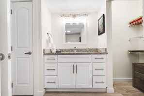 A bathroom with white cabinetry, a granite countertop, a mirror with lights, and a towel rack. - Sandcastle Condominiums Unit 10810 (Port Aransas)