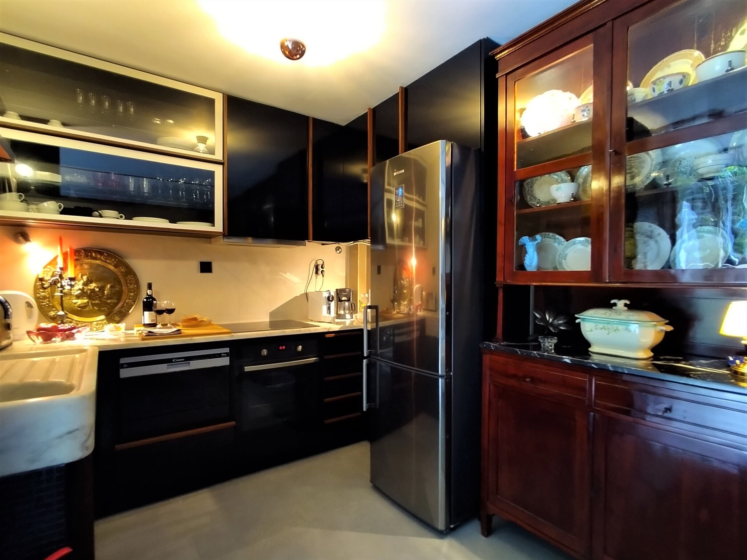 A modern kitchen with black cabinets, a stainless steel refrigerator, and a wooden cabinet with glass doors displaying dishes.