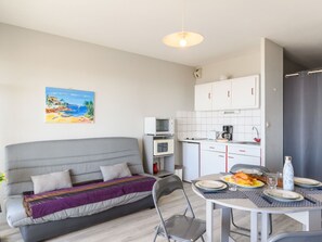 Un espacio habitable compacto con un sofá gris, una mesa de comedor para dos personas, una zona de cocina con armarios blancos y un cuadro con motivos playeros en la pared.