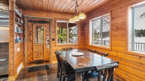 A wooden cabin interior with a dining table, benches, a kitchen area with a refrigerator, and large windows offering views of trees and a snowy landscape.