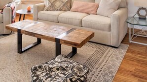 Ein Wohnzimmer mit einem beigefarbenen Sofa, einem gemusterten Hocker und einem Holztisch.