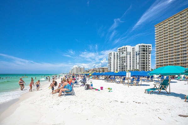 A beach with white sand, clear blue water, and tall buildings in the background.