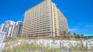 A high-rise hotel with multiple balconies, located near a beach with sparse vegetation and a clear blue sky. - Beautiful Updated Pelican Beach Unit!!! (Destin)