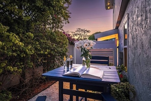 An outdoor patio with a table set for two, a book, a vase with flowers, and a chair.