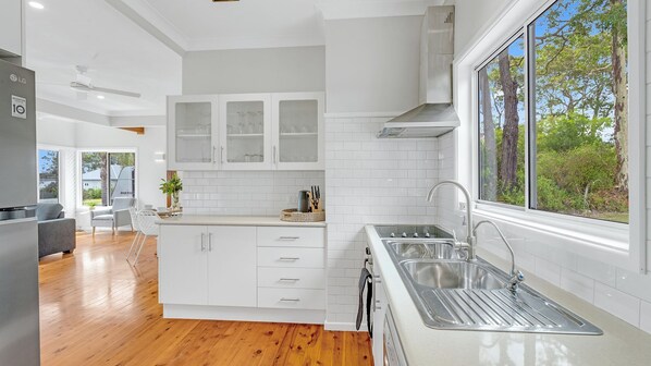 A modern kitchen with white cabinetry, a stainless steel sink, and a view of the outdoors through large windows. - Hazel's Haven, Erowal Bay (Erowal Bay)
