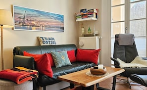 Un salón con un sofá de cuero negro, cojines rojos y azules, una mesa de centro de madera con una vela, una estantería llena de libros y un cuadro con una vista de la ciudad.