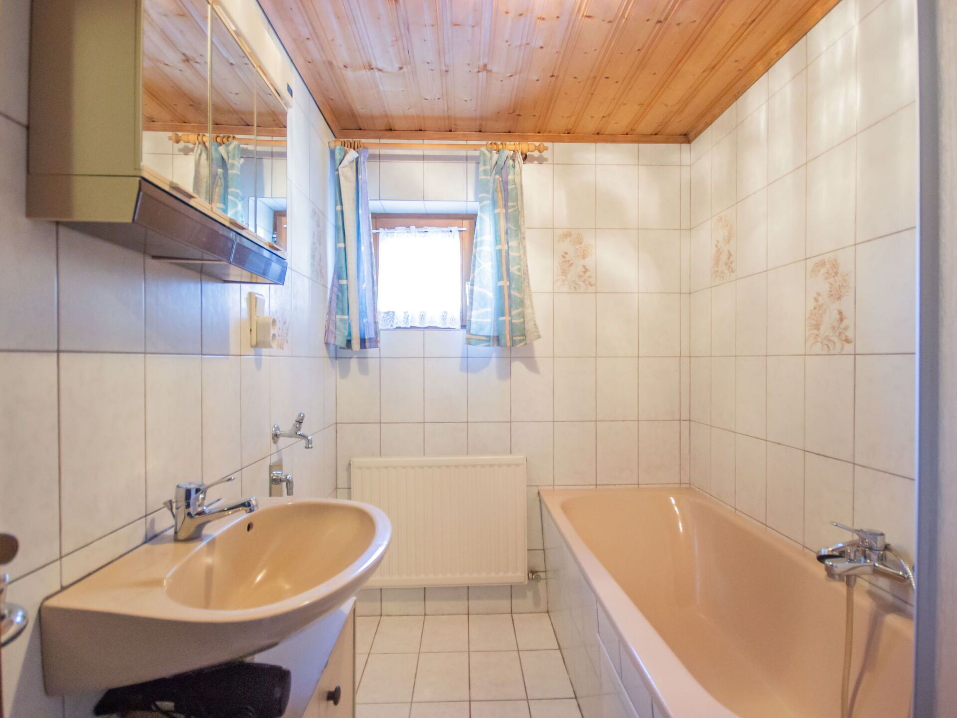 A bathroom with a wooden ceiling, white tiled walls, a bathtub, a sink, and a window with curtains.