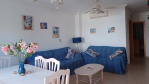 A living room with a blue sofa, white chairs, a small table, and a ceiling fan. - Captivating 3 bedroom House Casa Susan (San Juan de los Terreros)