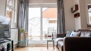 A living room with a sofa, a glass coffee table, a TV, and a large window with curtains. - Bedford Road Garden Apartment (Bedford)
