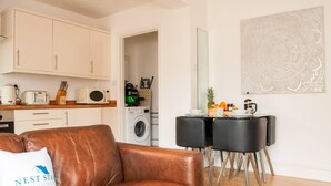 A compact living space with a brown leather sofa, a kitchenette with a microwave and toaster, a washing machine, and a dining area with two black chairs and a table. - Bedford Road Garden Apartment (Bedford)