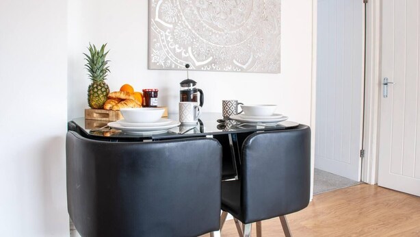 A dining area with two black chairs, a table with a coffee maker, a bowl, a cup, and a fruit basket. - Bedford Road Garden Apartment (Bedford)